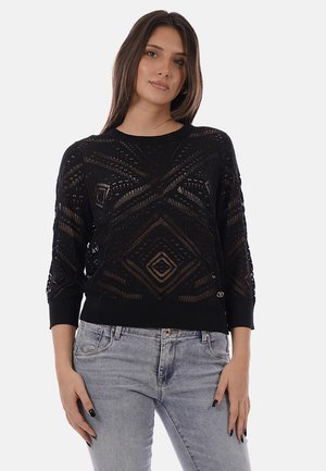 Donna che indossa un maglione nero a maniche lunghe in maglia aderente trasparente con motivi geometrici e jeans grigio chiaro su uno sfondo semplice.