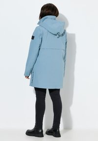 Chaqueta impermeable azul claro con capucha, cremalleras laterales y detalle negro en la manga; presenta un ajuste holgado y una textura suave.