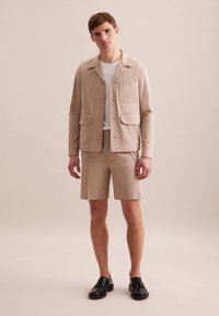 Giacca leggera beige con quattro tasche, indossata sopra una camicia bianca e pantaloni corti beige, abbinata a scarpe nere con lacci. Texture liscia.