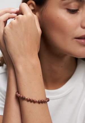 Frau trägt ein Perlenarmband mit rötlich-braunen und goldenen Perlen am Handgelenk, richtet ihr Haar und trägt ein weißes Hemd.