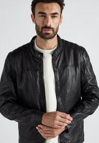 Mann, der eine schwarze Lederjacke über einem weißen Hemd trägt, steht vor einem einfarbigen hellen Hintergrund und zupft seinen Ärmel.