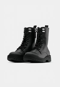 Crne gležnjače s vezicama od glatkog sintetičkog materijala. Imaju masivne đonove, srebrne metalne ukrase i prednji patentni zatvarač.