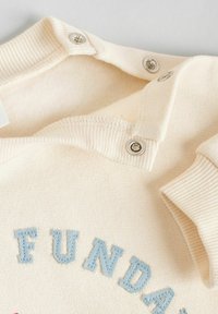 Cremfarbener Stoff mit weicher Textur, auf dem blaue gestickte Buchstaben zu sehen sind. Druckknöpfe am Kragen sorgen für Funktionalität.