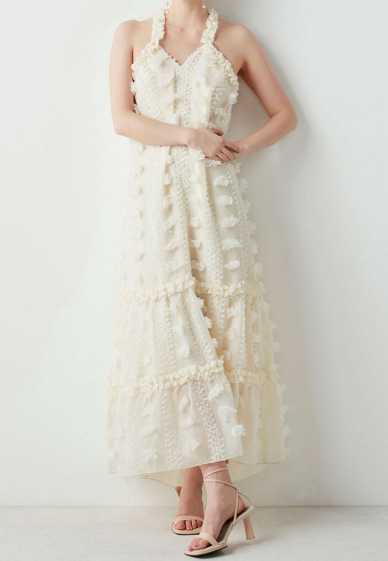 Femme portant une robe longue sans manches de couleur crème, avec une texture et des détails à volants, ainsi que des sandales beiges à talons hauts et à brides.