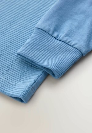 Une chemise à manches longues bleu clair présente une texture côtelée et des poignets élastiques, mettant en valeur un tissu léger et lisse avec des coutures soignées.