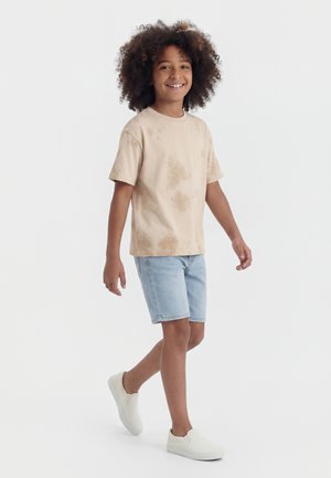 Niño con pelo rizado camina sonriendo, lleva una camiseta beige teñida anudada, pantalones cortos de mezclilla azul claro y zapatos blancos sin cordones.