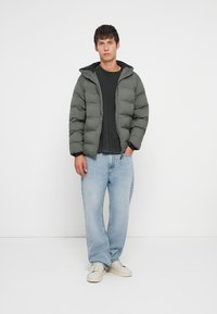 Πράσινο μπουφάν με φερμουάρ, ρυθμιζόμενη κουκούλα και ελαστικούς καρπούς, φορεμένο πάνω από μαύρο πουκάμισο και ανοιχτό μπλε τζιν, επιπλέον με λευκά αθλητικά παπούτσια.