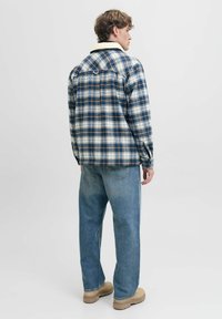 Chemise en flanelle à carreaux bleus, blancs et beiges avec un col en polaire. La personne qui la porte a desjeans bleu clair et des chaussures beiges.