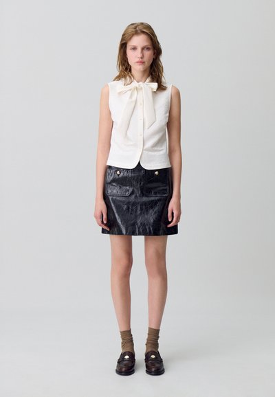 Mujer de pie con blusa blanca sin mangas con un gran lazo, falda corta de cuero negra con bolsillos frontales, calcetines marrones y mocasines oscuros.