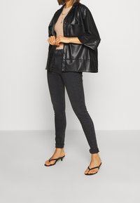 Camisa chaqueta oversized de cuero sintético negro, jeans oscuros ajustados y sandalias negras de tiras. Los detalles notables incluyen un bolsillo en el pecho y mangas sueltas.