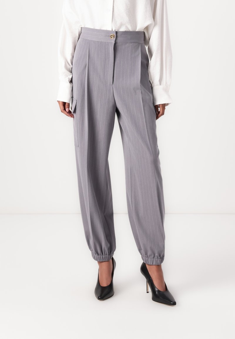 Pantalon gris à rayures avec une taille haute, fermeture à bouton à l'avant et poignets élastiqués ; associé à des chaussures à talons hauts noires.