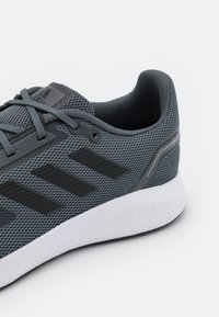 Šedá sportovní bota s texturovaným síťovaným svrškem, černými pruhy a bílou žebrovanou podrážkou. Má šedé tkaničky a doplňky.