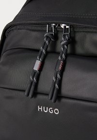 Crna ruksak od izdržljivog materijala s patentnim zatvaračem, pletenim trakama za povlačenje i istaknutim srebrnim logotipom "HUGO".