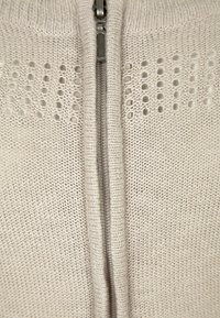 Maglione grigio chiaro lavorato a maglia con zip frontale, caratterizzato da piccoli motivi circolari intagliati vicino alle spalle e un tessuto testurizzato.