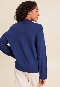 Pull en tricot bleu marine avec un col haut, des épaules tombantes, des poignets et un ourlet côtelés. Tissu texturé avec une coupe décontractée.