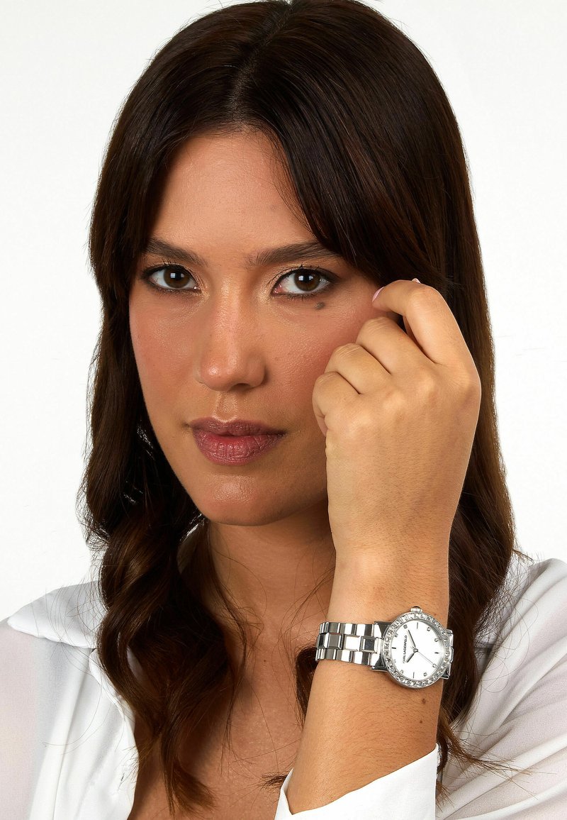 Runde silberne Armbanduhr mit weißem Zifferblatt, metallischem Armband und Strassakzenten auf der Lünette. Modell hält das Handgelenk nahe dem Gesicht.
