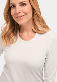 Witte lange mouwen shirt met ronde halslijn en een getextureerde trim. Model met licht golvend haar, neutrale achtergrond.