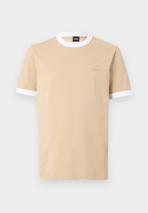To modeller iført beige t-shirts med hvide kanter; kvinde holder sort skuldertaske, mand bærer beige kasket, sorte korte bukser og loafers med hvide sokker.