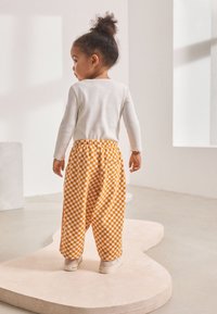 Chemise blanche à manches longues, pantalon à carreaux orange et jaune, coupe ample, texture douce, associé à des chaussures de couleur claire, vu de dos.