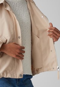 Giacca leggera beige con bottoni a pressione e chiusura con zip, interno foderato, indossata sopra un maglione in maglia bianco, con una tasca laterale visibile.