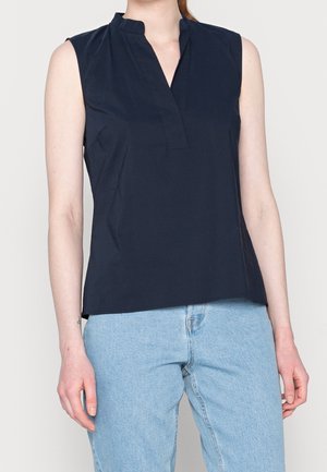 Haut bleu marine sans manches avec col en V porté avec un jean bleu clair, montrant le haut du torse et les bras sur un fond uni.