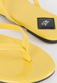 Infradito gialli con cinturini lucidi e lisci e una patch con logo nera. Hanno una suola piatta e un design minimalista.