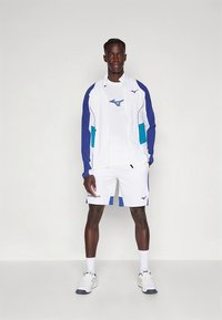 Mizuno sportkleding met een wit jack met blauwe en groene accenten, gecombineerd met een wit t-shirt en korte broek, allemaal gemaakt van lichtgewicht materiaal.
