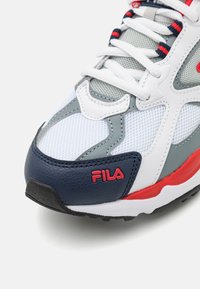 Białe i szare sneakersy z czerwonymi akcentami. Wykonane z materiałów siateczkowych i skórzanych, posiadają teksturowaną podeszwę oraz wyraźne logo Fila z boku.