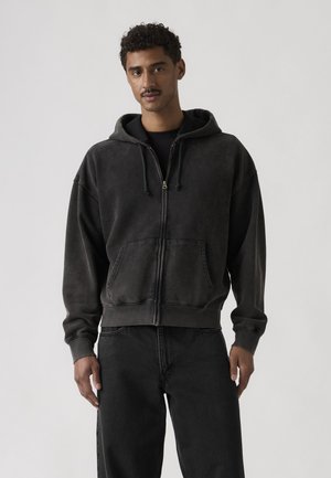 Hombre con una sudadera con capucha gris oscuro con cremallera y bolsillos frontales, y pantalones negros, posando frente a un fondo claro y sencillo.