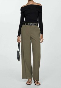 Vrouw in zwarte off-shoulder top, olijfgroene wijde broek met zwarte riem, draagt een kleine zwarte handtas en zwarte hoge sandalen.