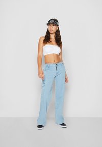 Un jean cargo bleu clair avec des poches à patch, associé à un haut blanc sans bretelles. Une casquette noire avec des détails en motif blanc vient compléter la tenue.