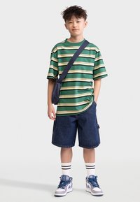 Green and yellow striped t-shirt, blue denim shorts, white socks with black stripes, gray and blue sneakers, and a black crossbody bag.