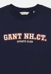 Tumši zils T-krekls, izgatavots no kokvilnas, ar baltu un sarkanu grafikā tekstu "GANT NH.CT. SPORTS CLUB" treknrakstā. Apaļa kakla izgriezuma forma.