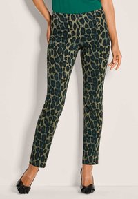Vrouw in slim-fit panterprint broek en zwarte pointed heels, staand tegen een lichte muur. 