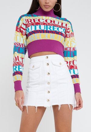 Kvinde iført en farverig cropped sweater med fed tekst og en hvid højhældet denim mininederdel med knapper.