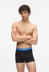 Jaunas vyras su garbanotais plaukais, dėvintis juodus apatinės kelnės su mėlynu dirželiu, pažymėtu "KARL LAGERFELD", stovi susikėlęs rankas.