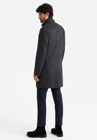 Abrigo gris de espiga con cuello alto, corte entallado, bolsillos laterales y pantalones oscuros contrastantes, combinado con botas de tobillo negras.