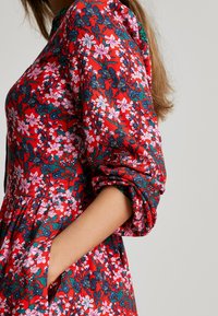 Czerwona sukienka w kwiaty z różowymi i ciemnoniebieskimi kwiatami, długie rękawy, marszczona faktura oraz boczne kieszenie. Posiada luźny krój i okrągły dekolt.