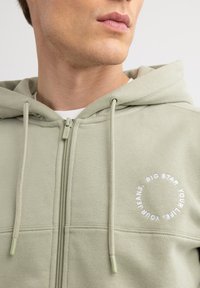Helle grüne Zip-Up-Hoodie aus weichem Stoff mit einem runden weißen Logo auf der linken Brust und passenden Kordelzügen.
