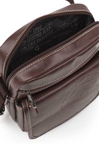Bolso cruzado de cuero marrón con dos compartimentos con cremallera, costuras blancas y un patrón de logo en relieve en la parte frontal, que incluye una correa desmontable.