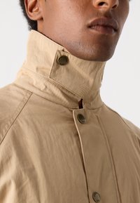 Beige jakke med høy krage, med en glatt tekstur, knappeåpning detaljer, og merkede knapper på kragen og forsiden.