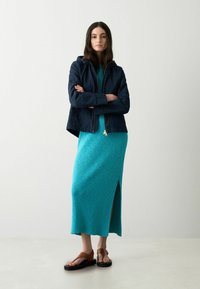 Gequilted marineblauwe jas met een rits en capuchon, gecombineerd met een teal ribgebreid jurkje met een zijsplit, en bruine sandaaltjes op een neutrale achtergrond.