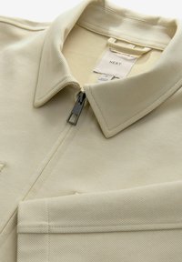 Bež jakna na zip s teksturiranim materijalom, okruglim ovratnikom i malom etiketom na izrezu. Sadrži metalni zip i šivene detalje.