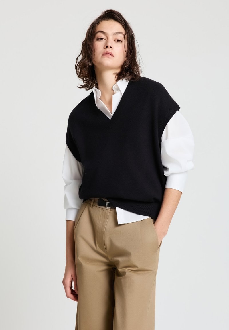 Schwarzer V-Ausschnitt-Pullover, der über einem weißen Hemd mit weiten Ärmeln getragen wird. Kombiniert mit beigen Hosen. Glatter Stoff und klassisches, minimalistisches Design.