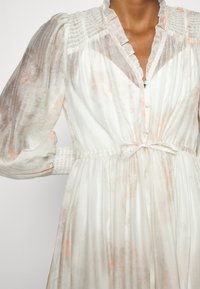 Robe blanche transparente avec un motif pêche subtil, manches longues bouffantes, épaules et poignets smockés, boutons à l'avant et un nœud à la taille.