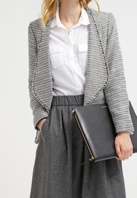 Blazer de espiga gris con un frente abierto, llevado sobre una camisa blanca. Combinado con una falda gris de cintura alta, sosteniendo un bolso de mano de piel negro.