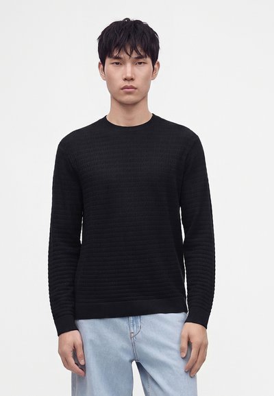 Homme portant un pull noir texturé à manches longues et un jean bleu clair debout devant un fond blanc uni.