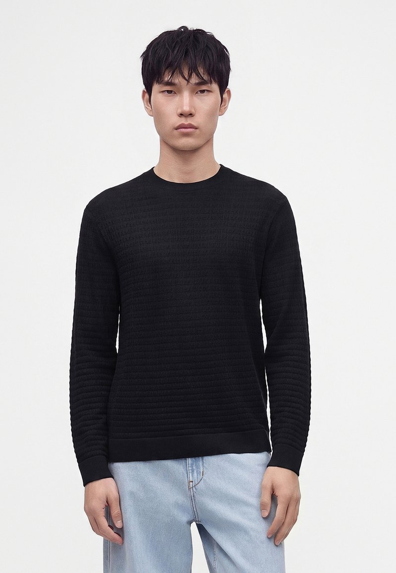 Homme portant un pull noir texturé à manches longues et un jean bleu clair debout devant un fond blanc uni.