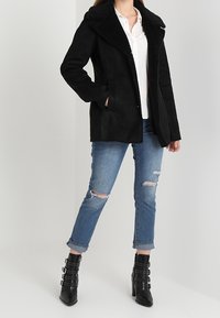 Czarna zamszowa kurtka z grubym, teksturowanym kołnierzem, dwoma bocznymi kieszeniami, połączona z przetartymi jasnoniebieskimi dżinsami i czarnymi botkami na obcasie.