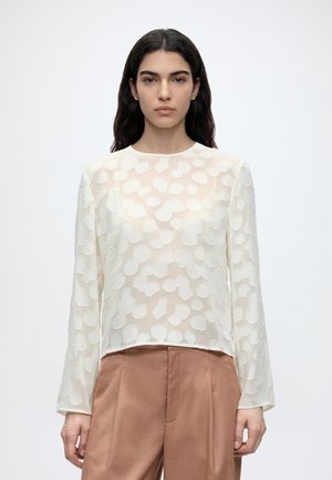 Femme aux longs cheveux foncés portant un chemisier blanc à manches longues à motifs et un pantalon marron taille haute, debout devant un fond uni.